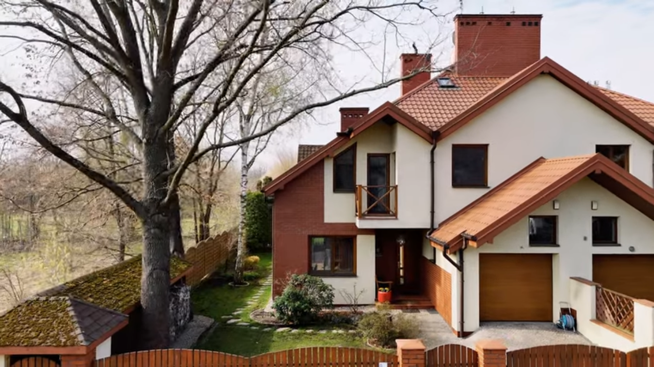 Nowoczesny dom jednorodzinny z czerwonym dachem, ogrodem i ogrodzeniem, w tle nagie drzewa.