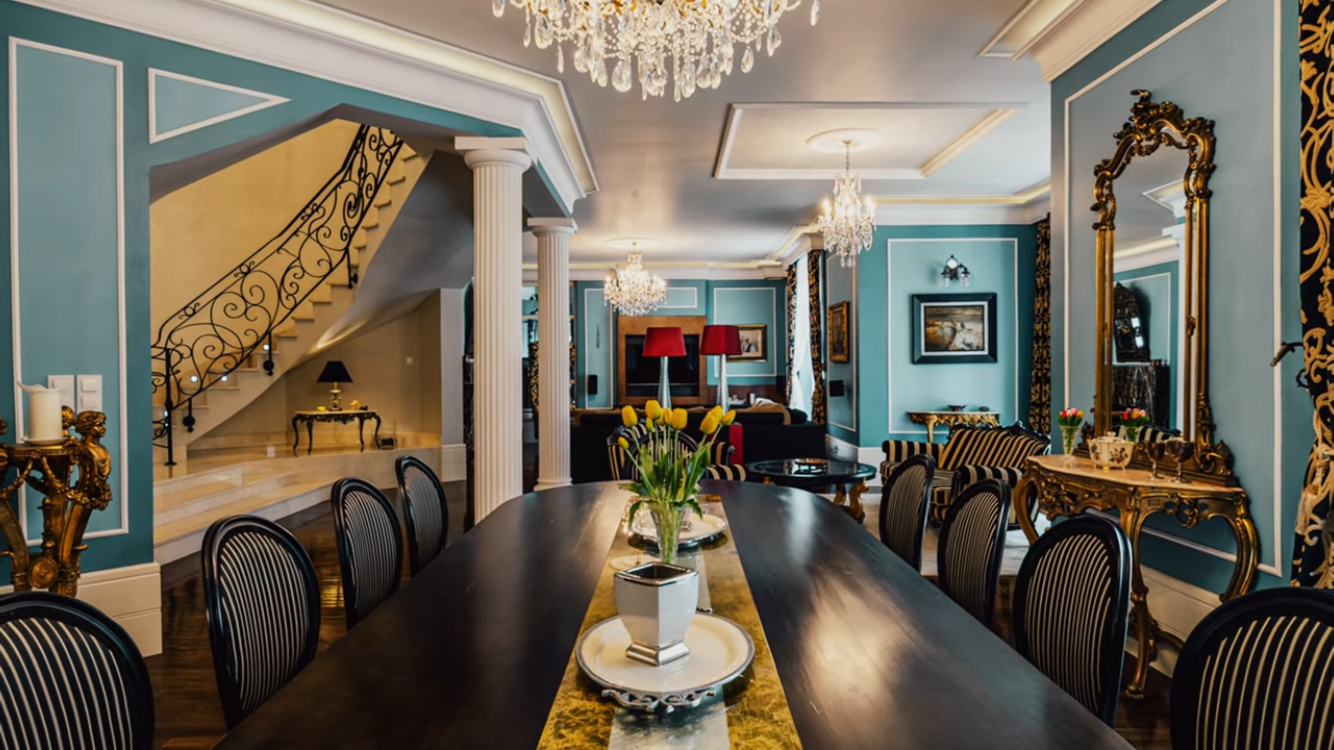 Elegant dining room with dark table, ornate chairs, chandeliers, teal walls, and gold-framed mirror.