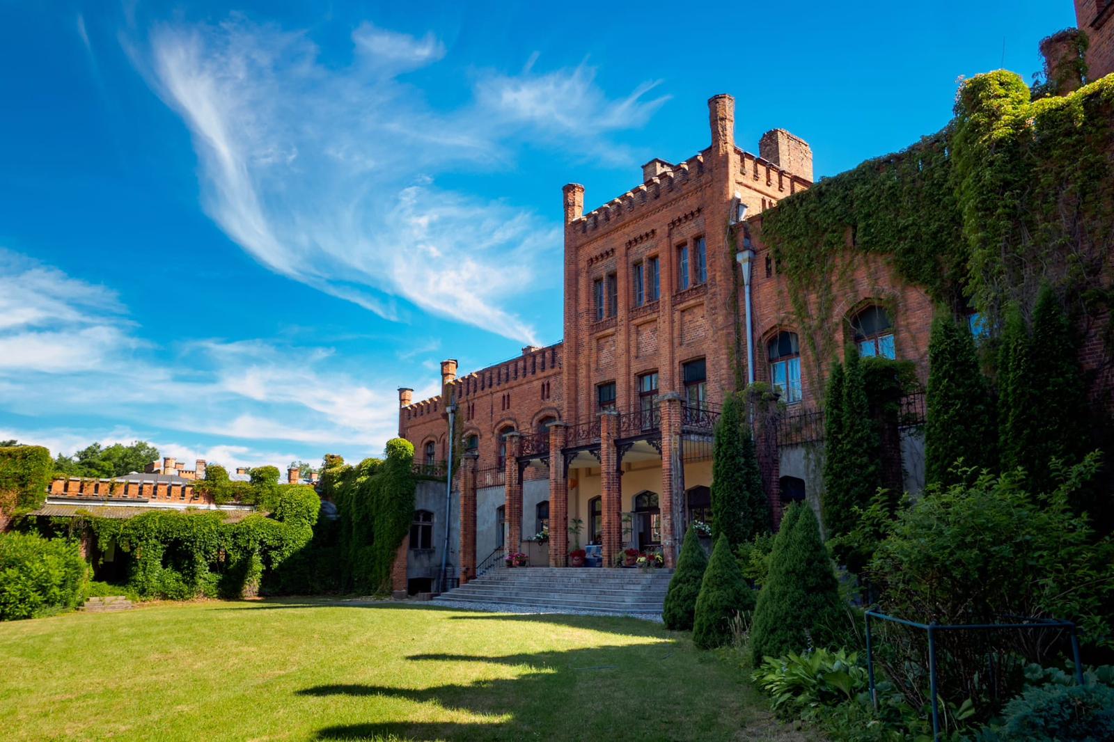 Historic red-brick building with tall chimneys, arched windows, and lush green gardens under blue sky.