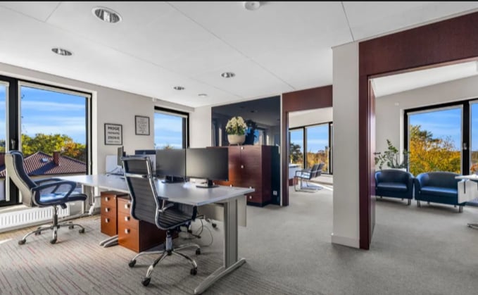 Modern office with desks, rolling chairs, glass partitions, and large windows overlooking trees and sky.