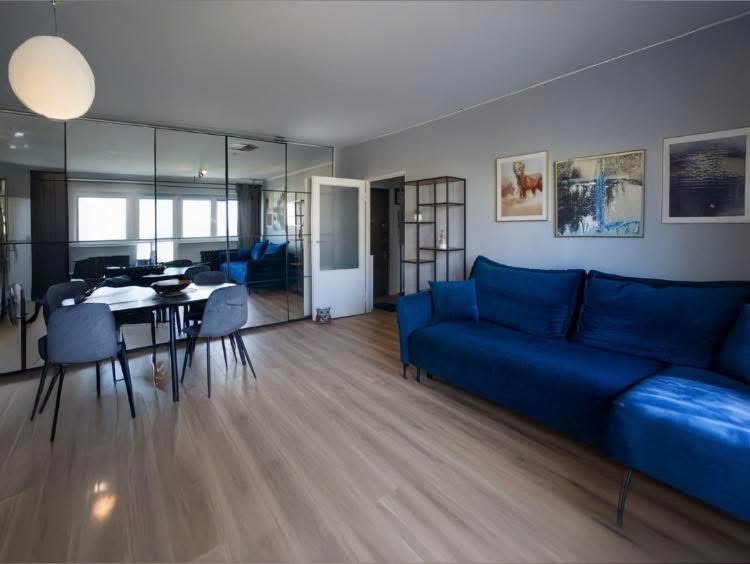 Bright modern living room with blue sofa, dining table, large windows, and framed wall art.