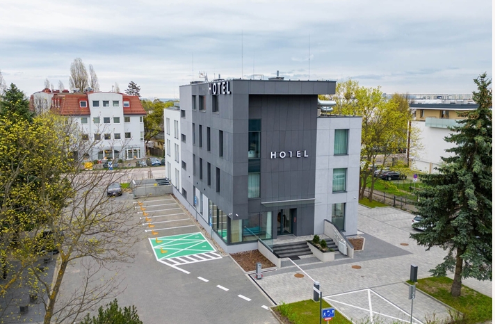 Nowoczesny hotel z napisem na budynku, parkingiem i oznakowanym placem, otoczony zielenią i drzewami.